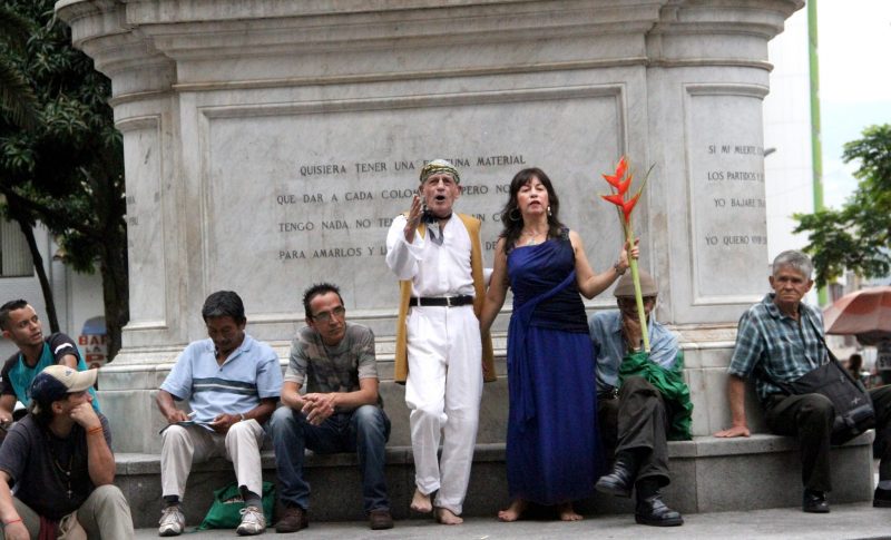 Two performers standing infront of a white marble monument with civilians sitting next to them.