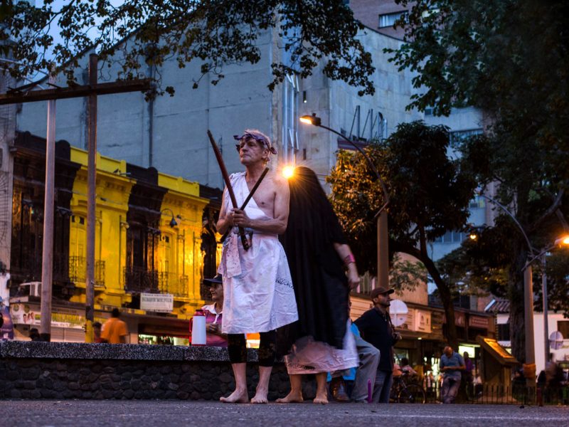 An older man in a toga stands barefoot in the street with two sticks in his hands.