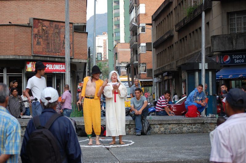 A man in yellow and a woman dressed as a nun perform in front of a street crowd.