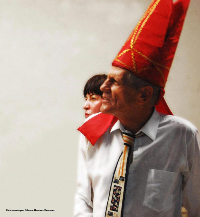 An older man with a large pointy red hat on looks off into the distance with a lady looking over his shoulder.