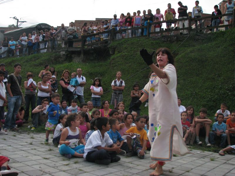 A woman performers in front of a group of children sitting on the ground with parents standing in the background.